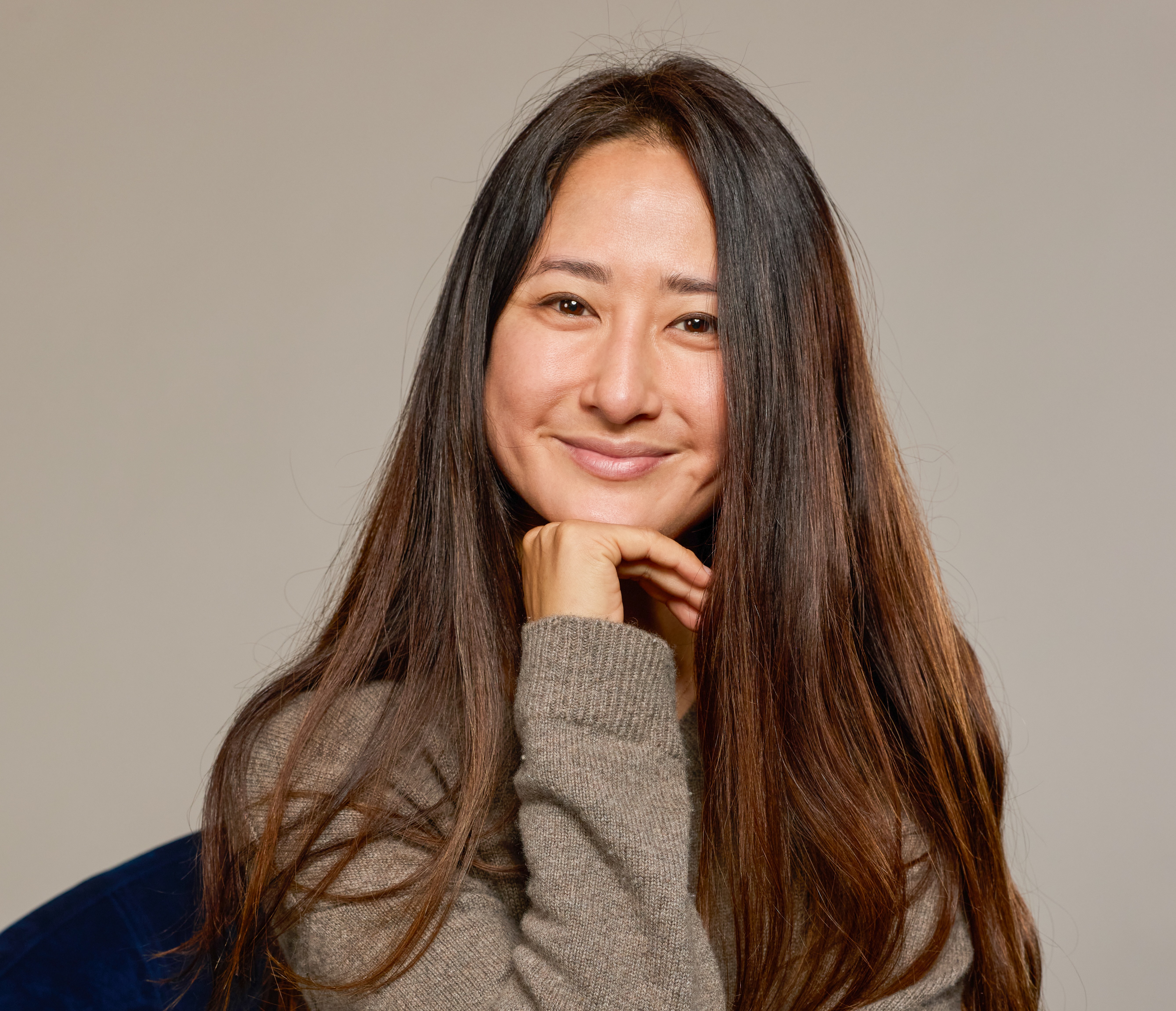 A smiling woman with long dark hair resting her chin on her hand, wearing a cozy sweater against a neutral background.